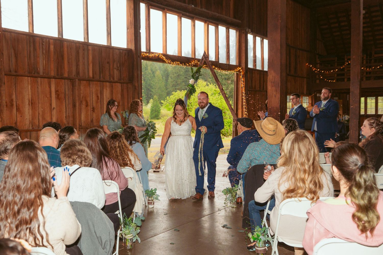 Long shot of wedding ceremony in wooden barn with natural lighting and gathered attendees.