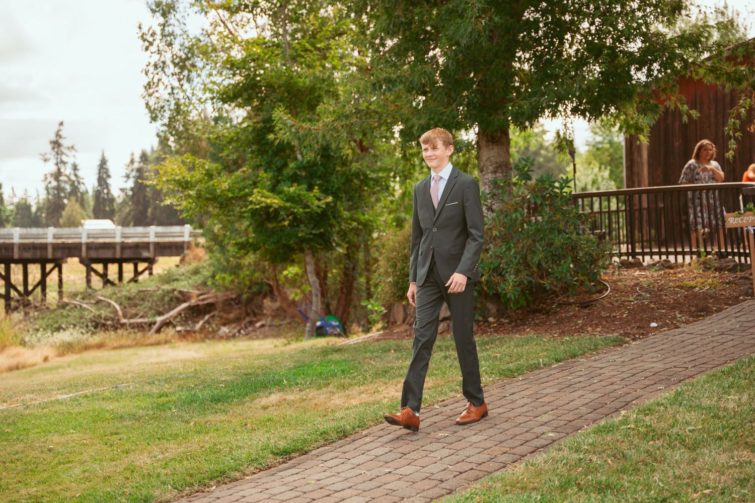 Person in dark suit walks along brick pathway surrounded by green lawn and trees.