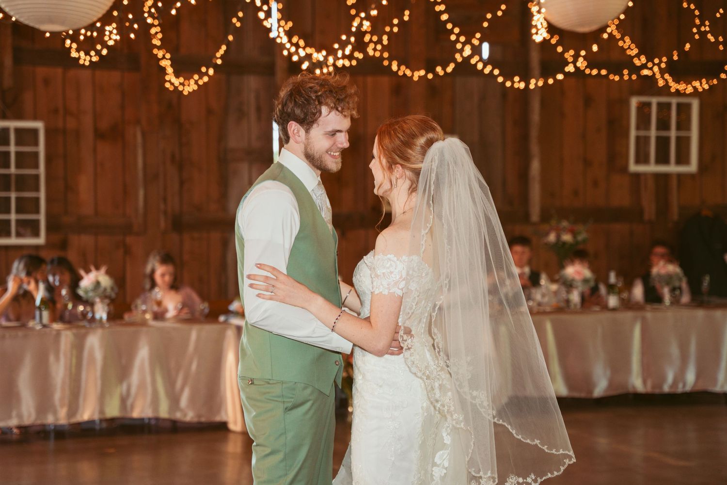 A couple shares their first dance in a rustic barn venue decorated with warm string lights and white decor.