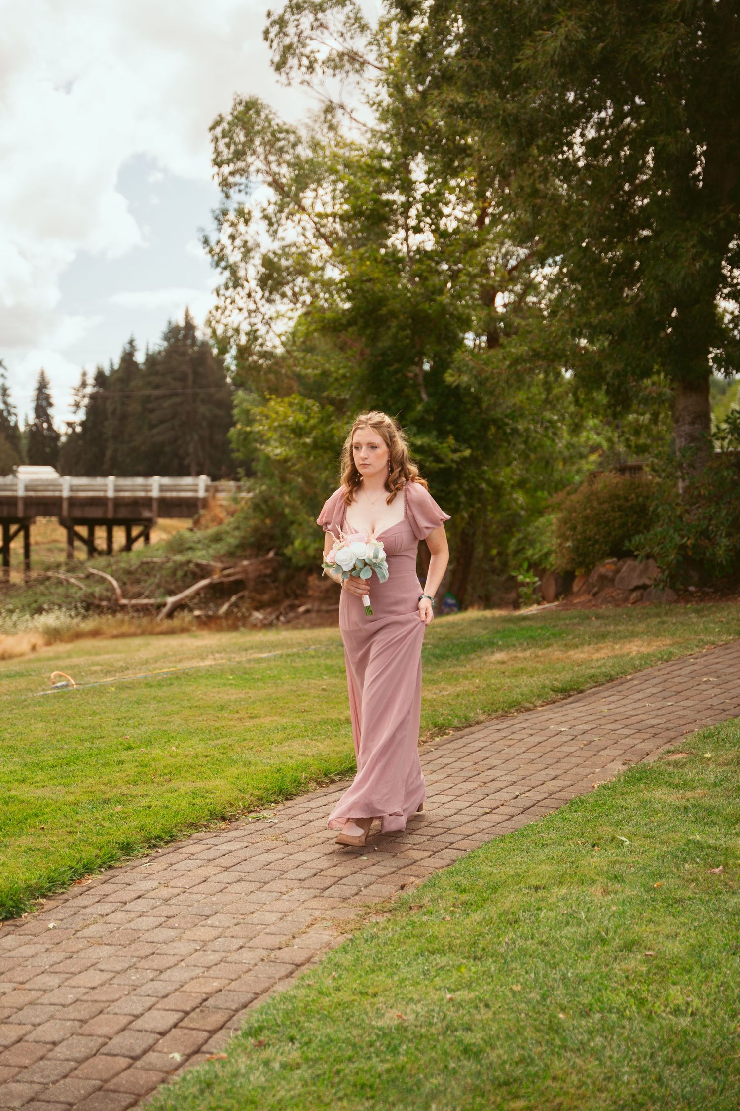 Someone in a pink gown walks along a winding brick path during an outdoor ceremony.