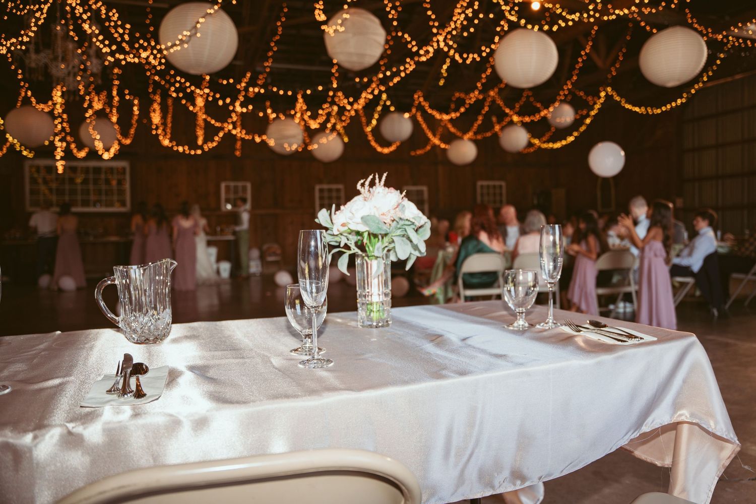 White-clothed tables set for a wedding reception under twinkling barn lights.