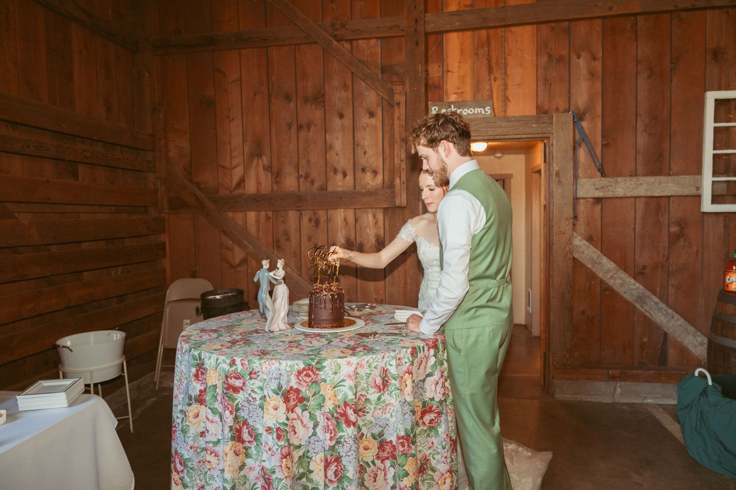 Wedding cake cutting preparation on floral tablecloth in rustic wooden barn interior.