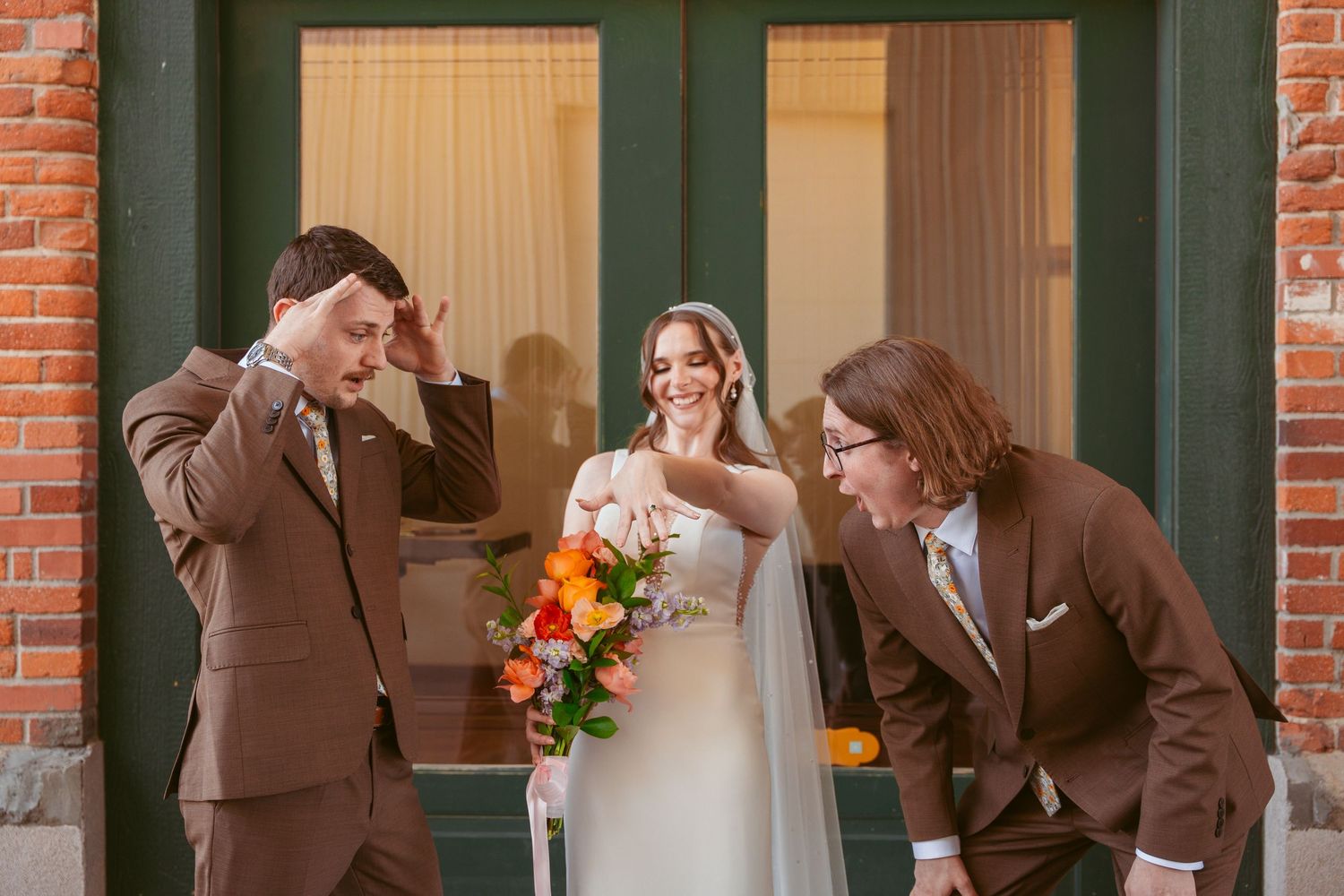 A fun wedding party photo with groomsmen in brown suits posing playfully outside a brick building.