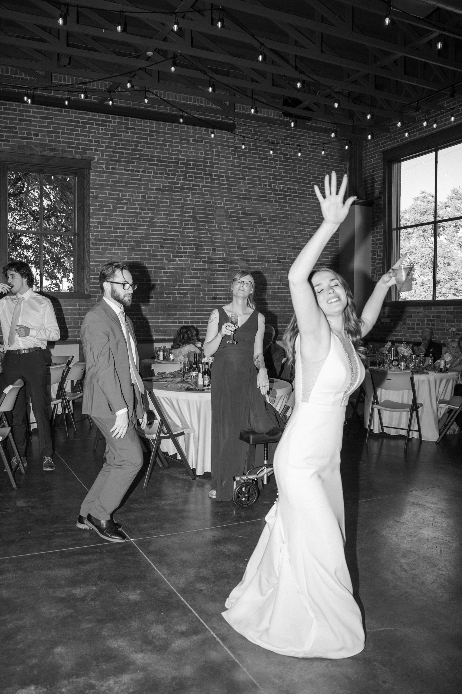 Black and white photo of wedding guests dancing joyfully under string lights in a brick venue.