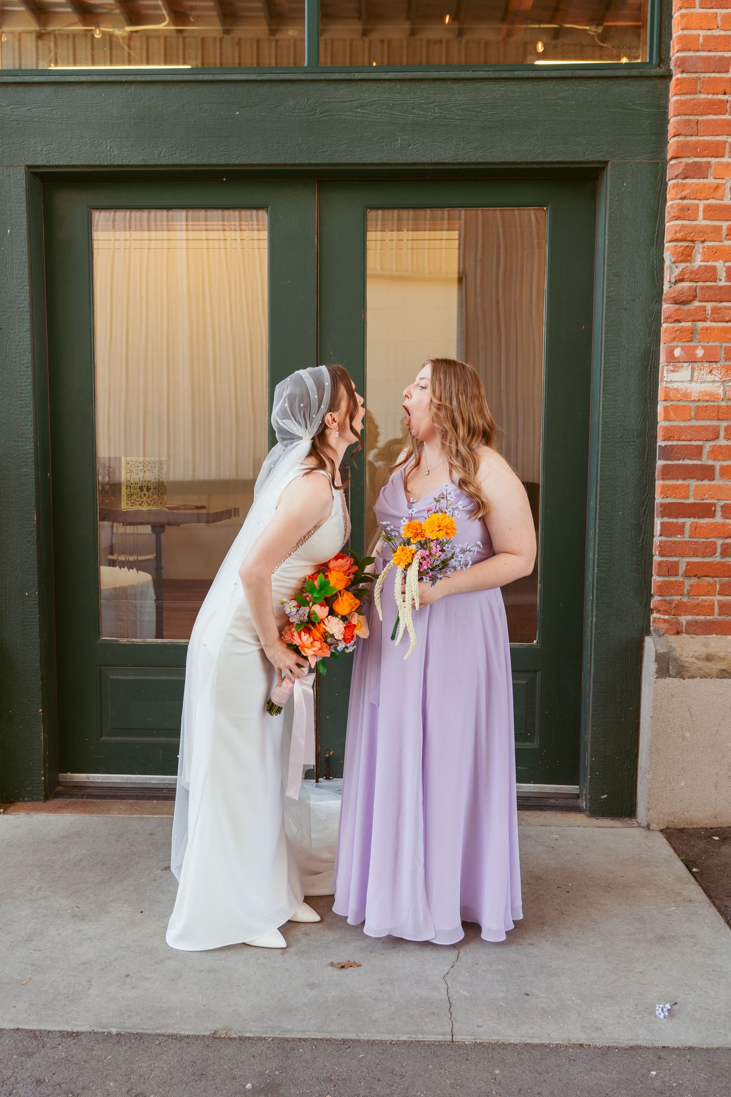 Bride in white dress and person in lavender gown share a moment by green doorway.
