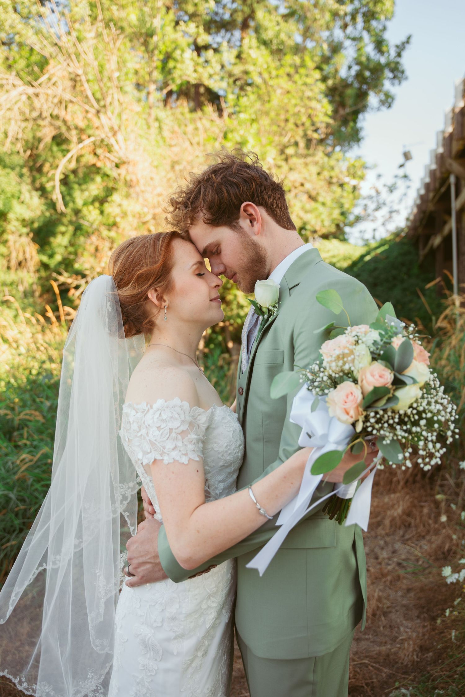 A romantic wedding moment captured outdoors with a sage green suit and lace dress with delicate florals.