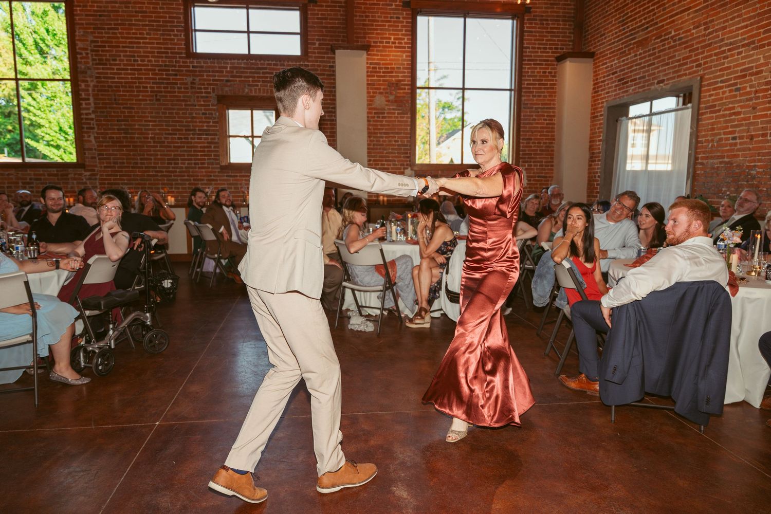 Wedding guests dance energetically in a rustic brick venue with large windows and natural lighting.