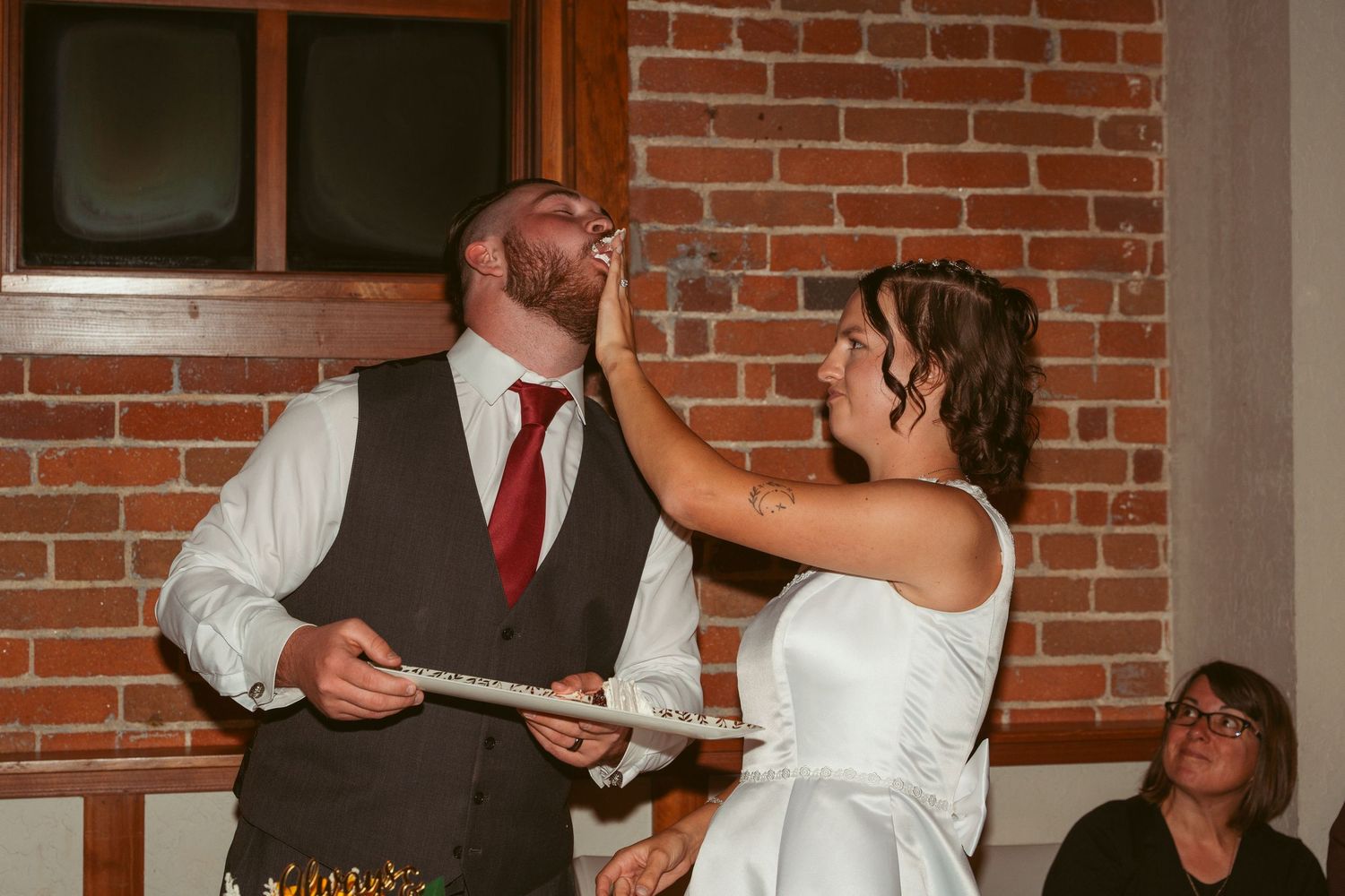 A playful cake-feeding moment between newlyweds at their wedding reception against a brick wall backdrop.