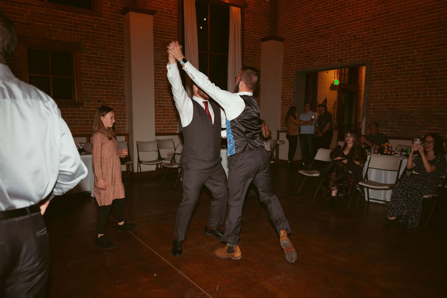 People dancing and celebrating at a wedding reception in a rustic brick-walled venue.