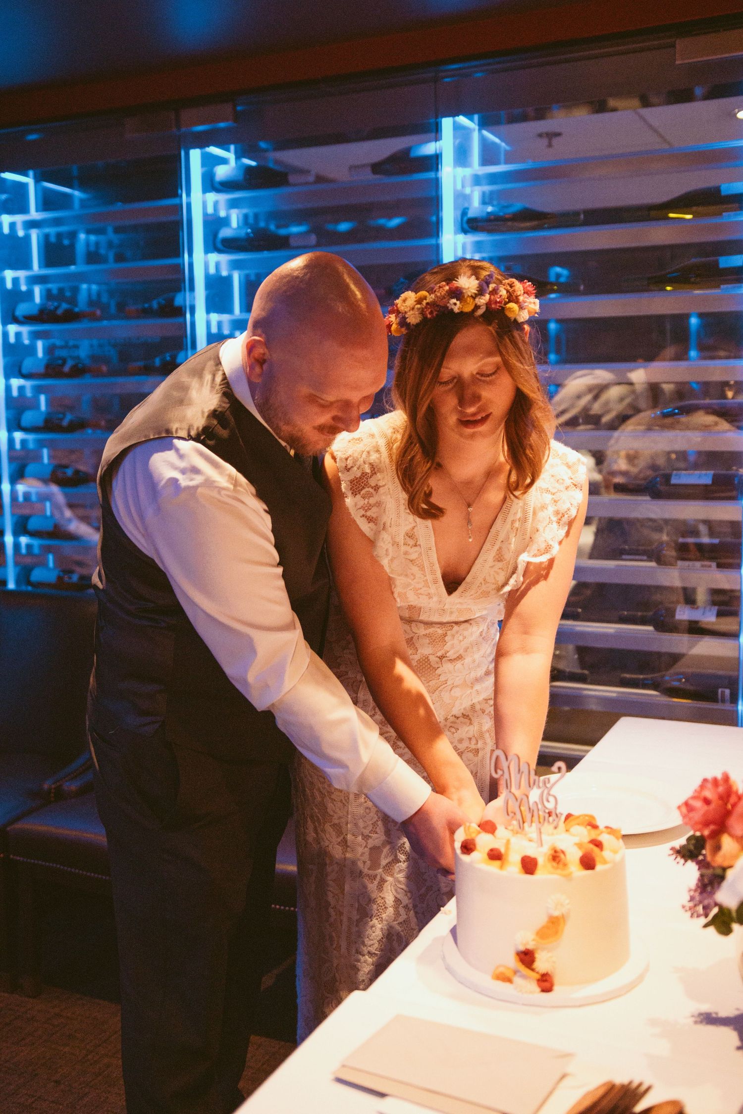 A couple cuts their white wedding cake together under blue ambient lighting.