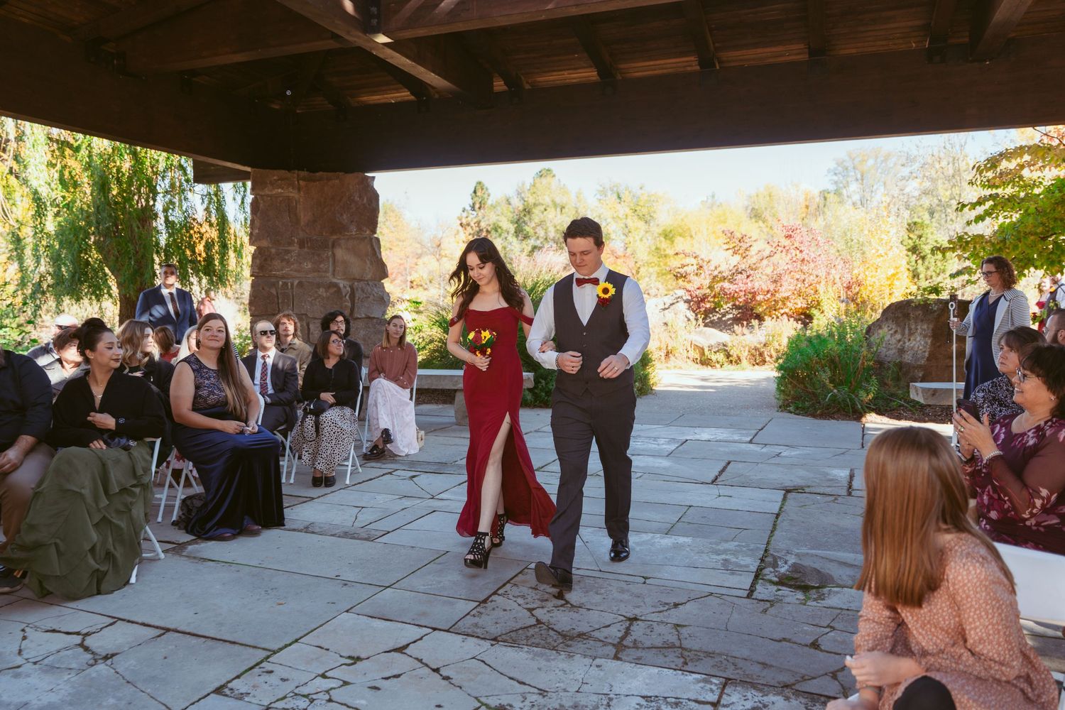 Wedding guests seated on benches during an outdoor ceremony under wooden pergola.
