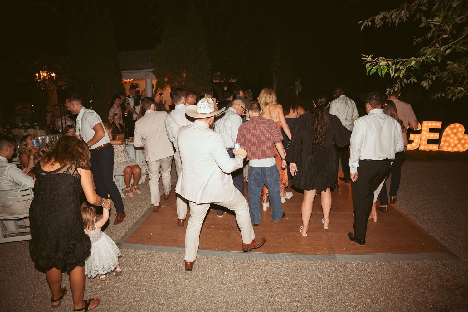 Guests dancing energetically at nighttime wedding reception with lit signage in background.