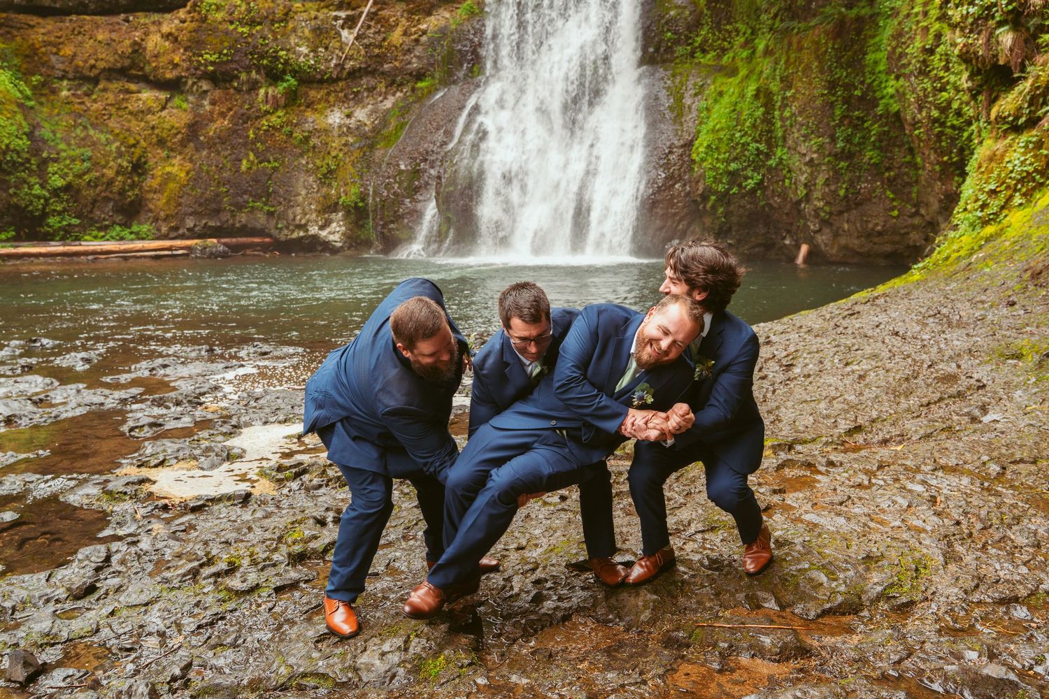 Wedding party members in navy suits crouch playfully by a forest waterfall.