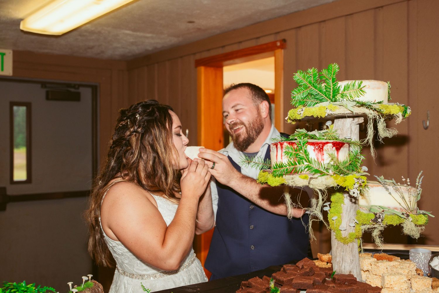 Wedding couple laughing while feeding each other cake at their indoor reception.