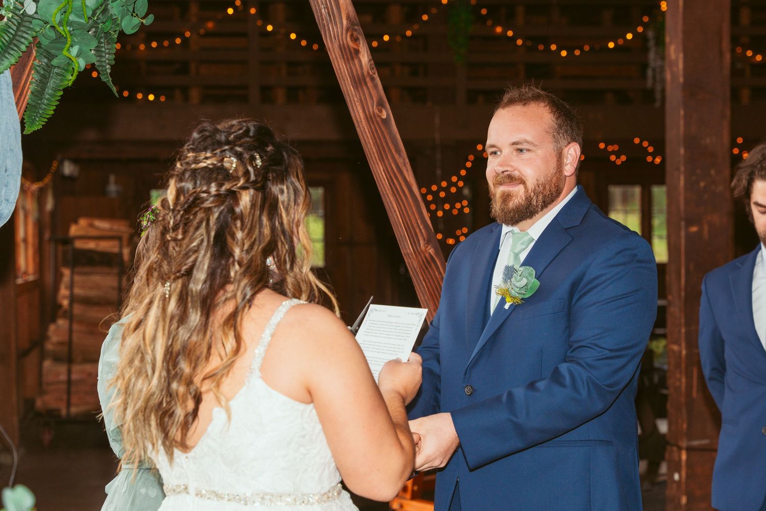 Romantic wedding ceremony moment in rustic barn with string lights and natural decor.