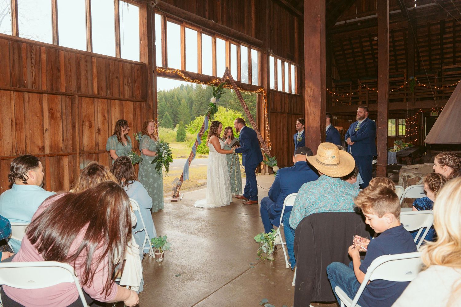 Interior view of a barn wedding ceremony with guests seated in folding chairs and natural light streaming in.