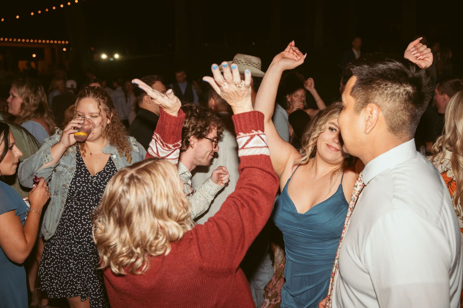 Large group of party guests dancing with raised arms under twinkling outdoor lights.
