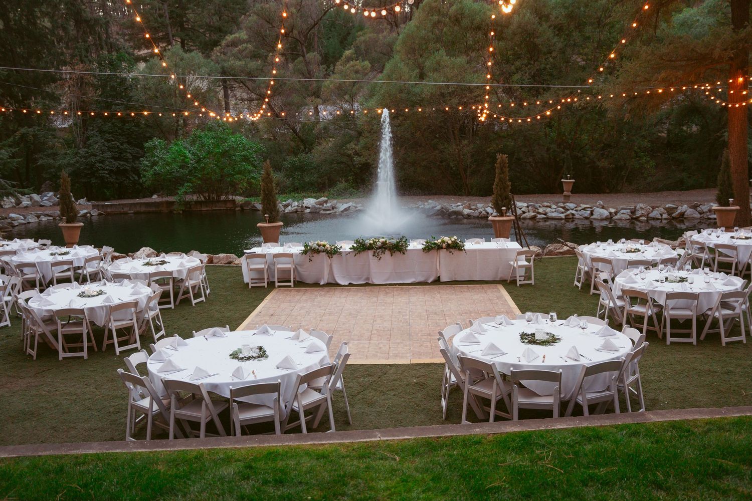 Elegant outdoor wedding reception setup with string lights and white tables arranged on a grassy lawn.