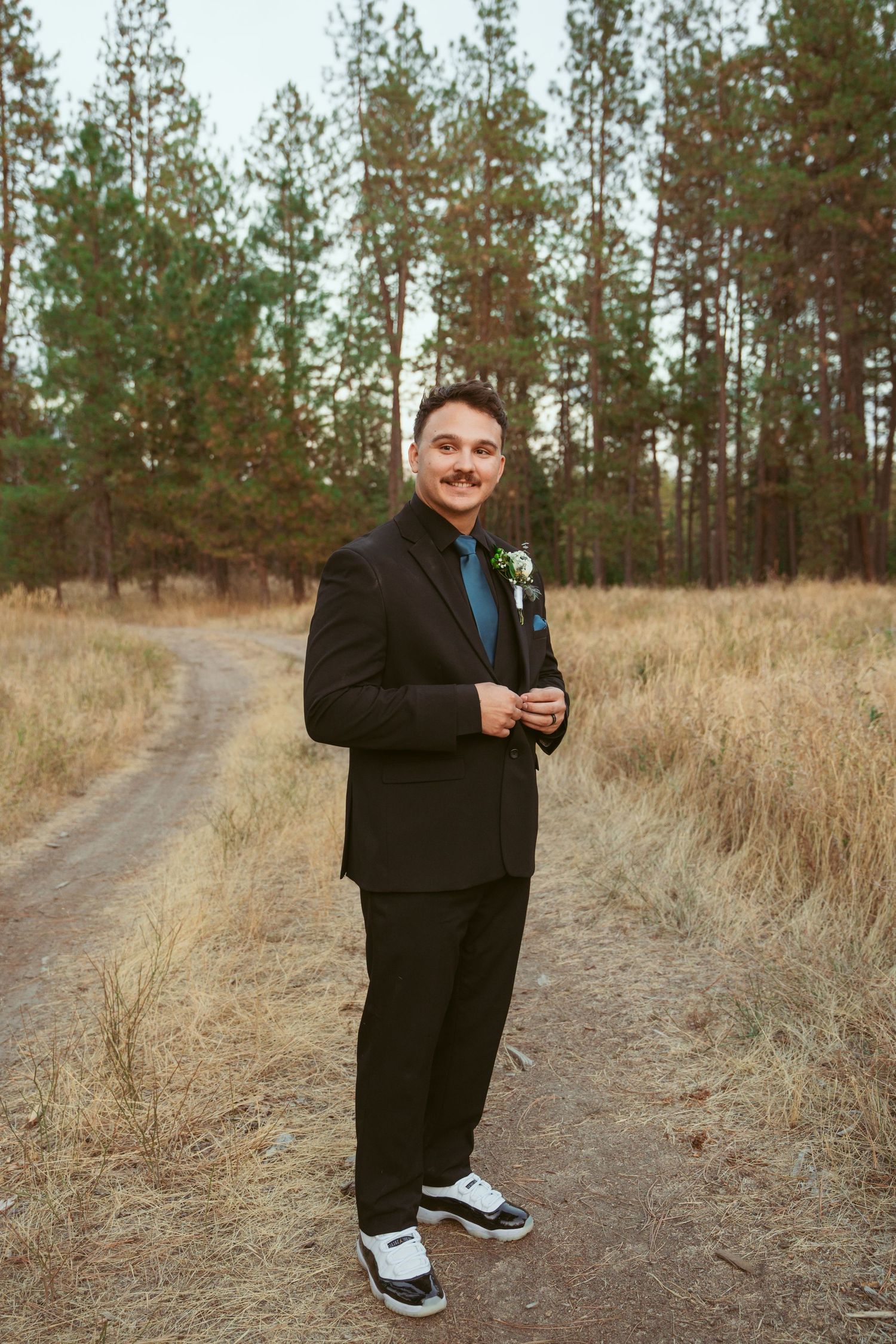 Groom in black suit with blue tie and sneakers poses playfully on dirt path surrounded by tall pine trees.