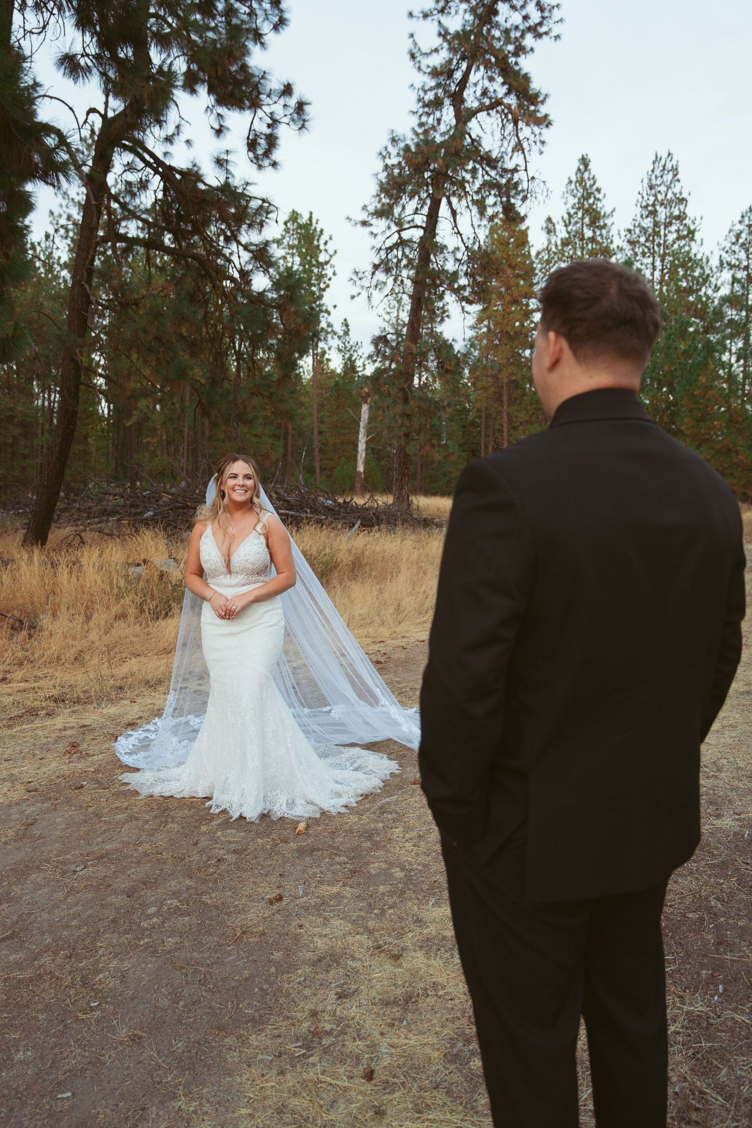 A couple shares a romantic first look moment on a dirt path surrounded by pine trees.