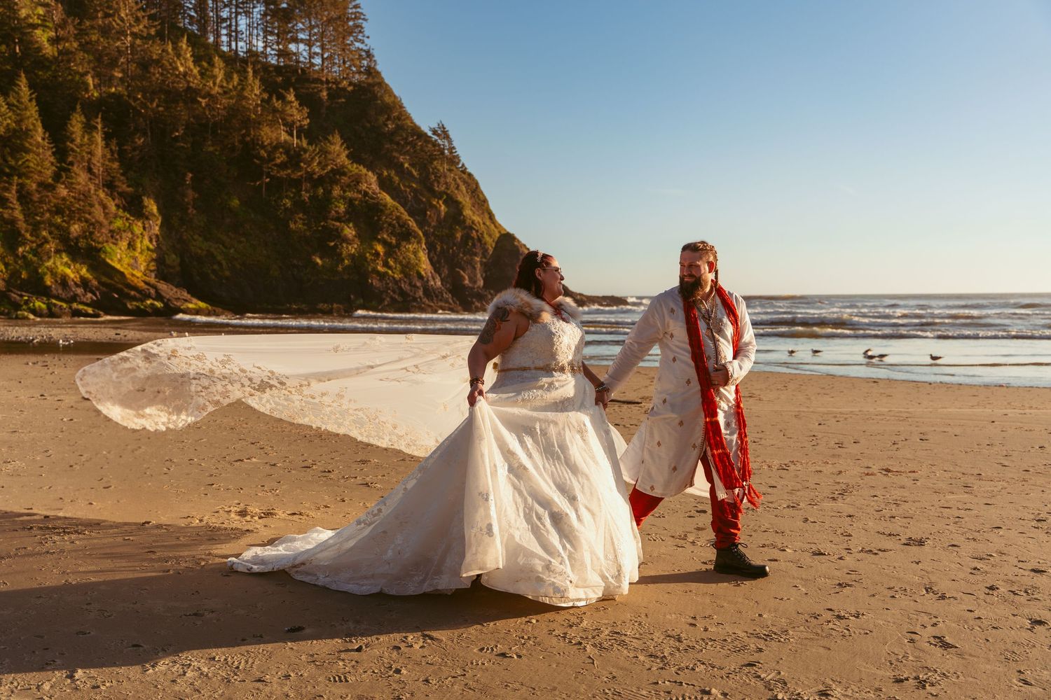 A couple poses playfully on the beach during golden hour with dramatic coastal cliffs behind them.