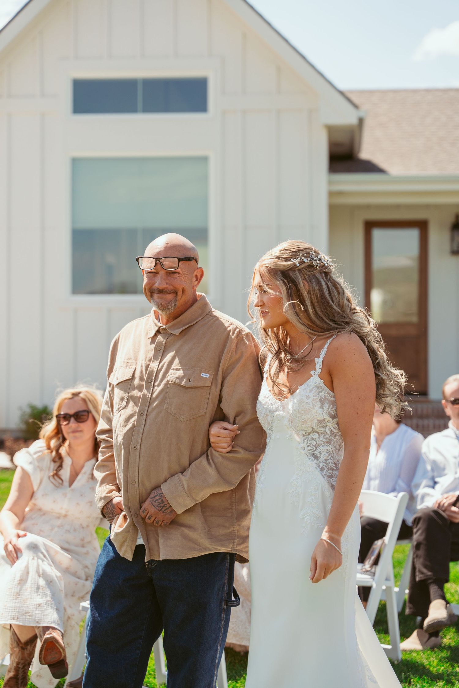 Wedding party walking together on grass outside white farmhouse-style building on sunny day.