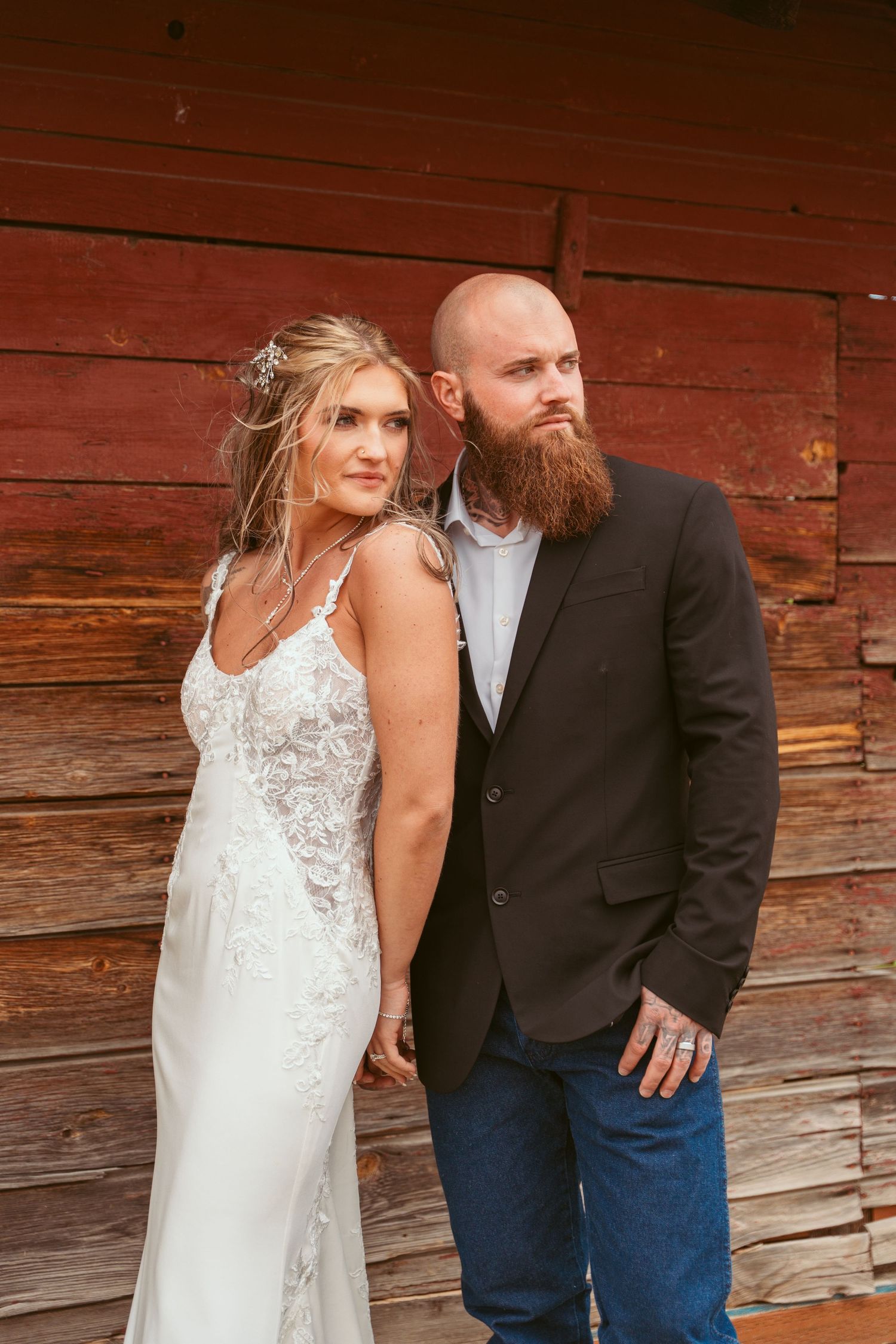 A couple poses against a rustic red barn wall in wedding attire.