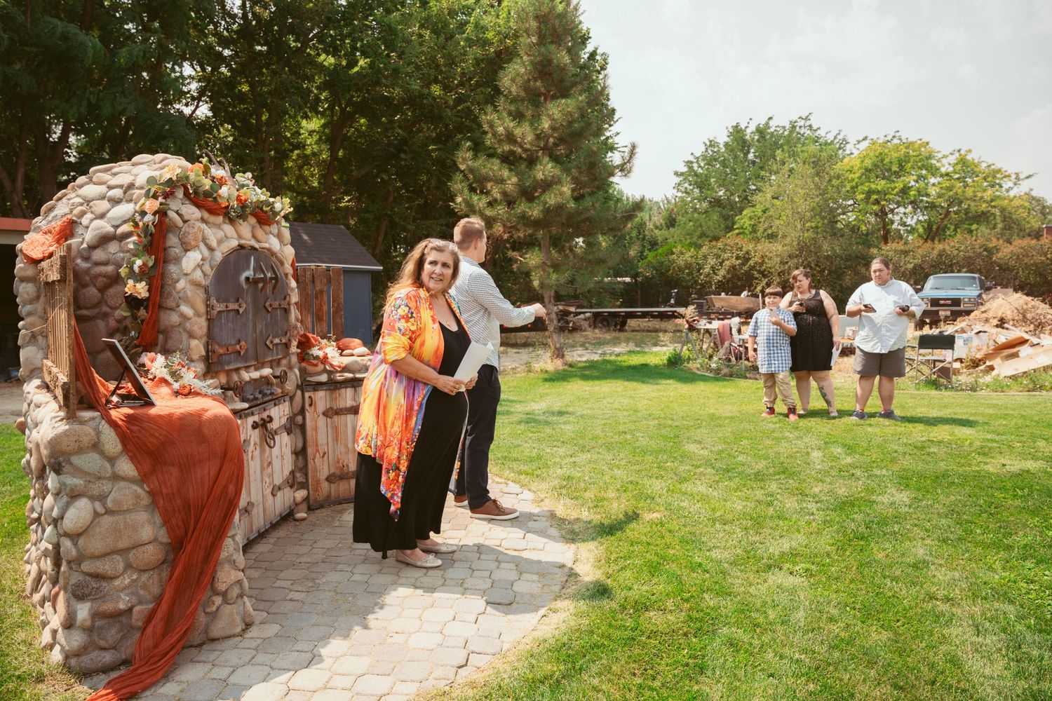 Wedding guests gather outdoors in a garden setting with stone walls and green lawn for a celebration.