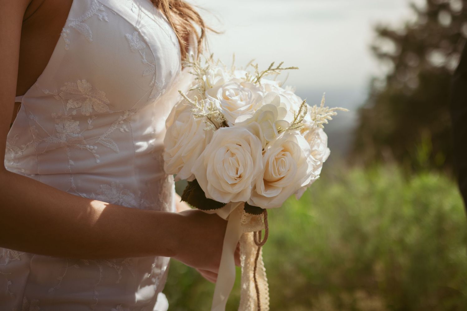 Close-up of a white wedding dress and bouquet details in natural sunlight.