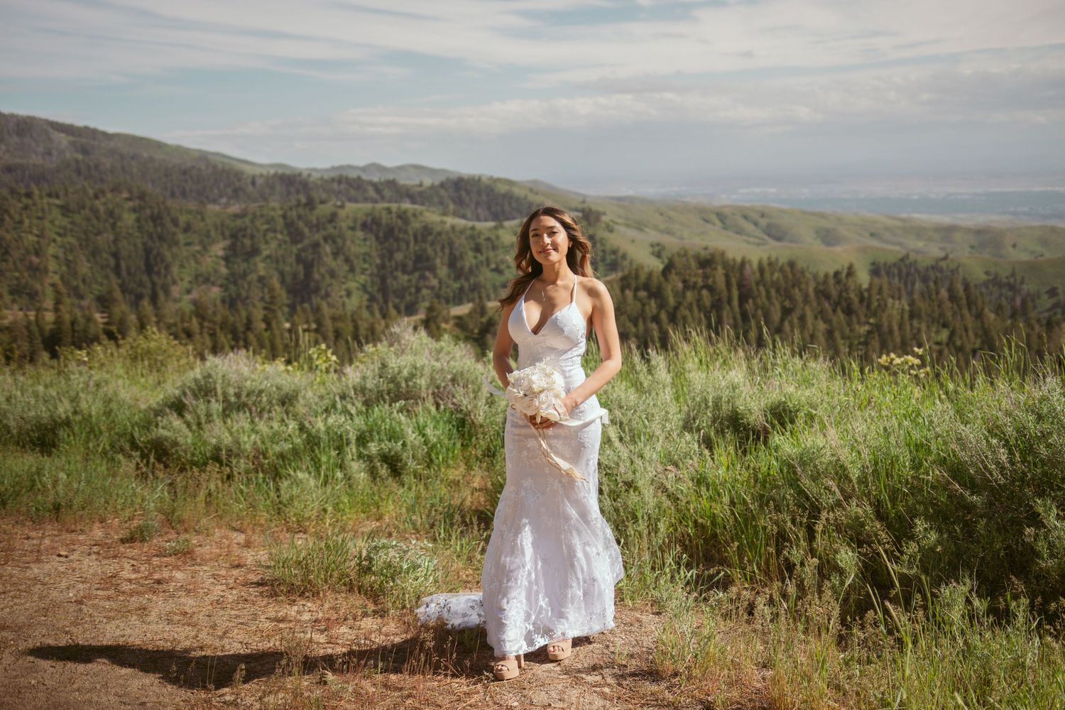 A bride in a white lace dress holds a bouquet while standing on a mountain trail with valley views.