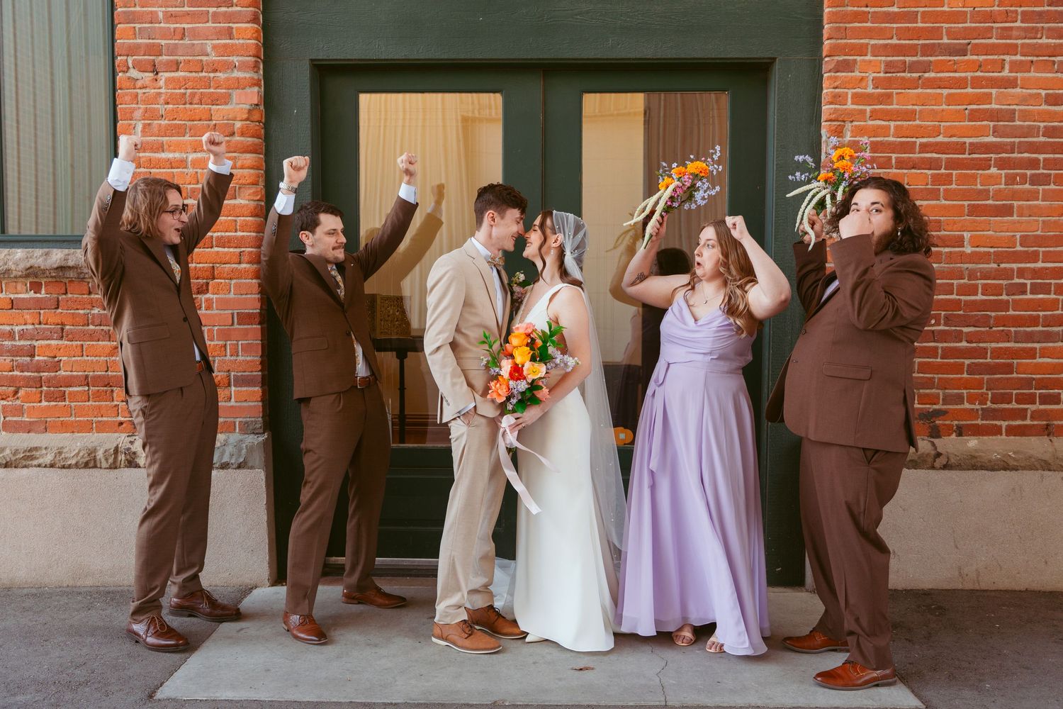 Bridal party in purple and brown attire posing casually against brick wall.