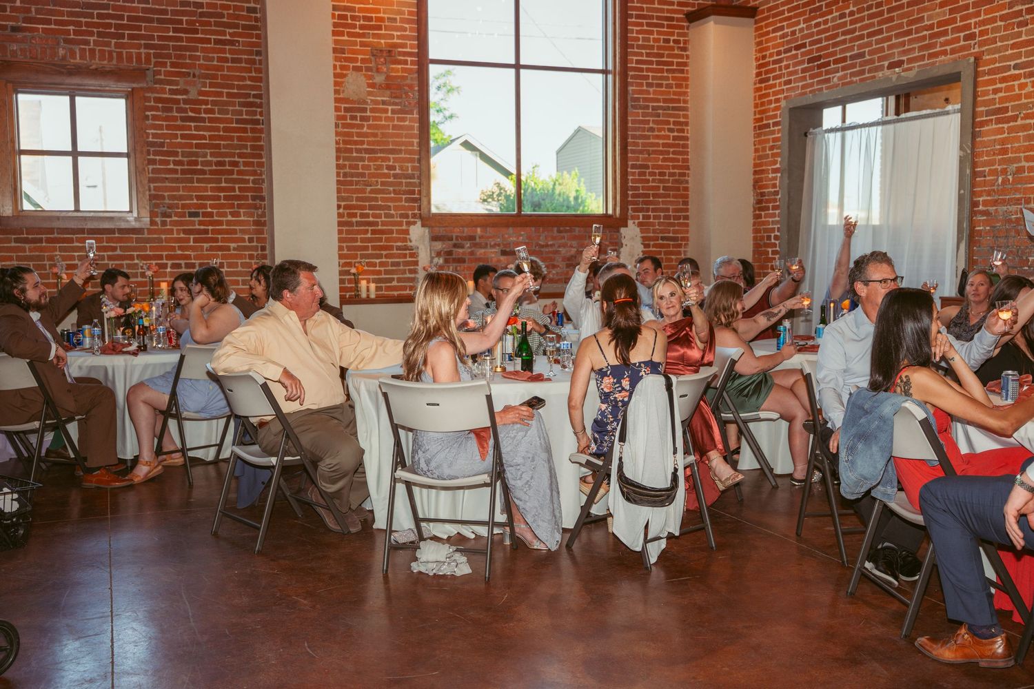 Wedding reception guests seated at tables in a rustic brick-walled venue with natural lighting.