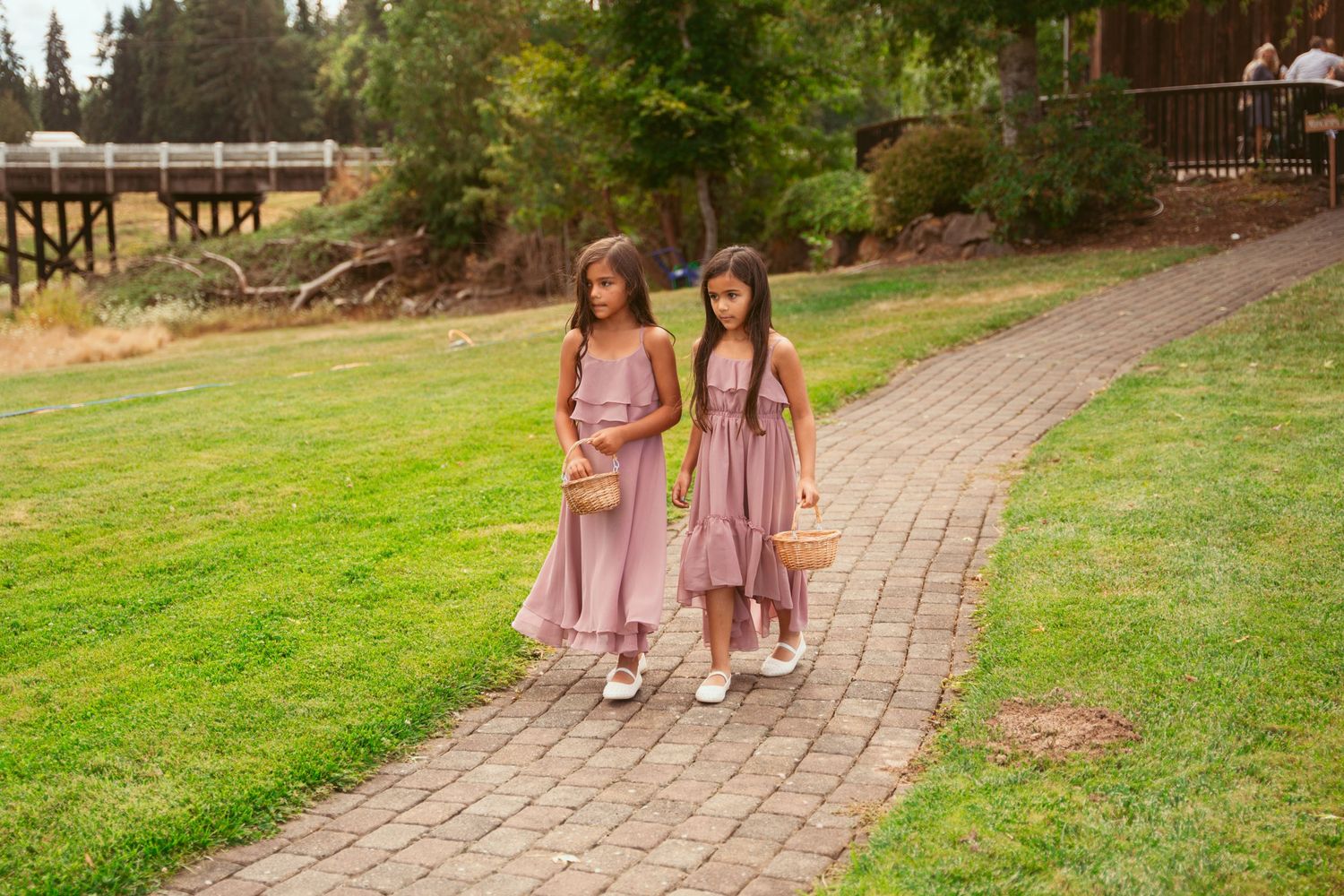 Young attendants in pink dresses walk down a brick pathway at an outdoor event.