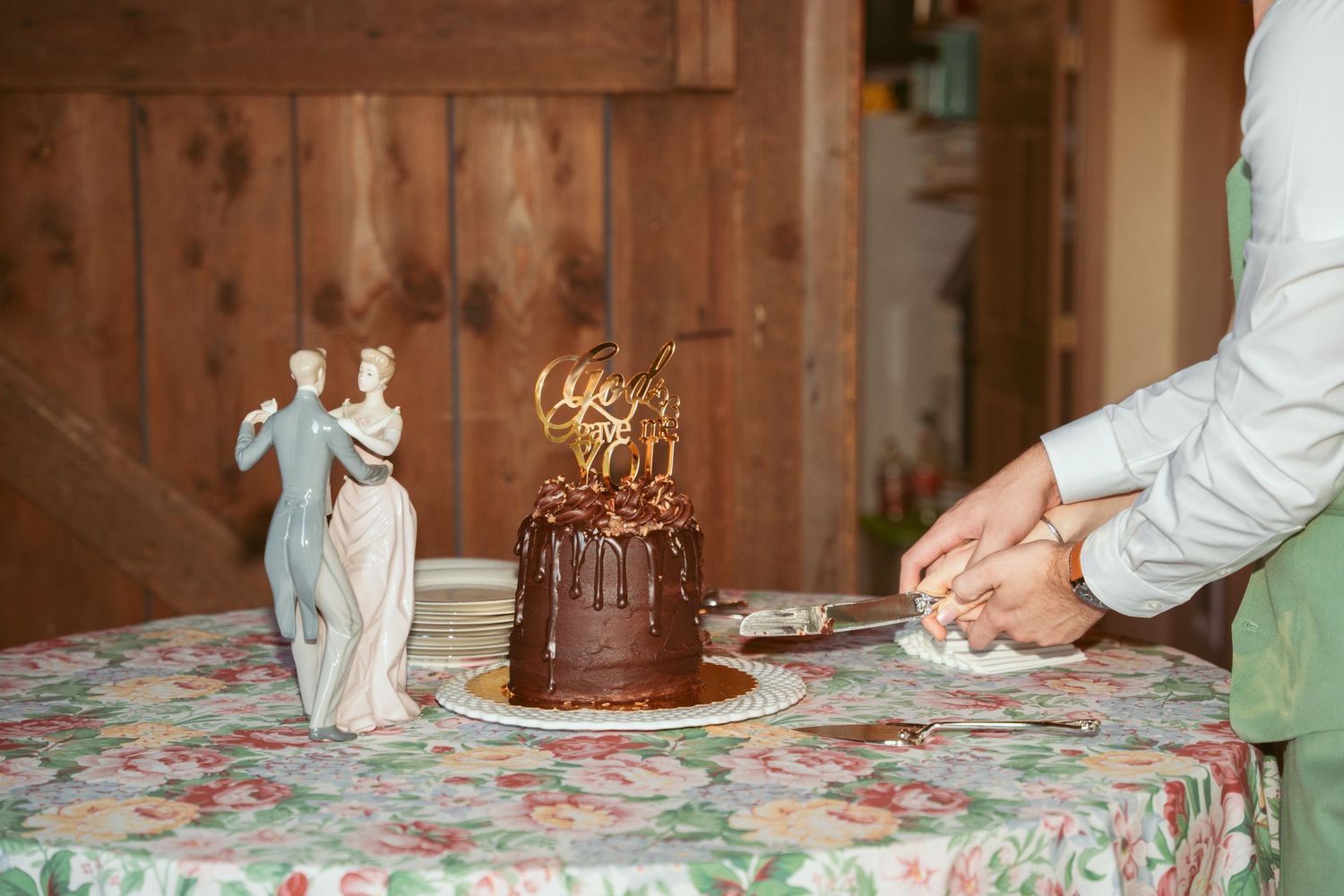 Chocolate wedding cake being cut on floral tablecloth at rustic reception.