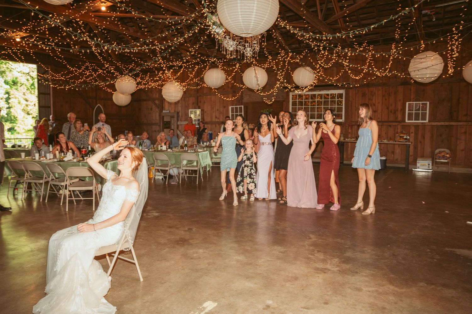 Wedding guests dance and celebrate under twinkling string lights and paper lanterns in a rustic barn venue.
