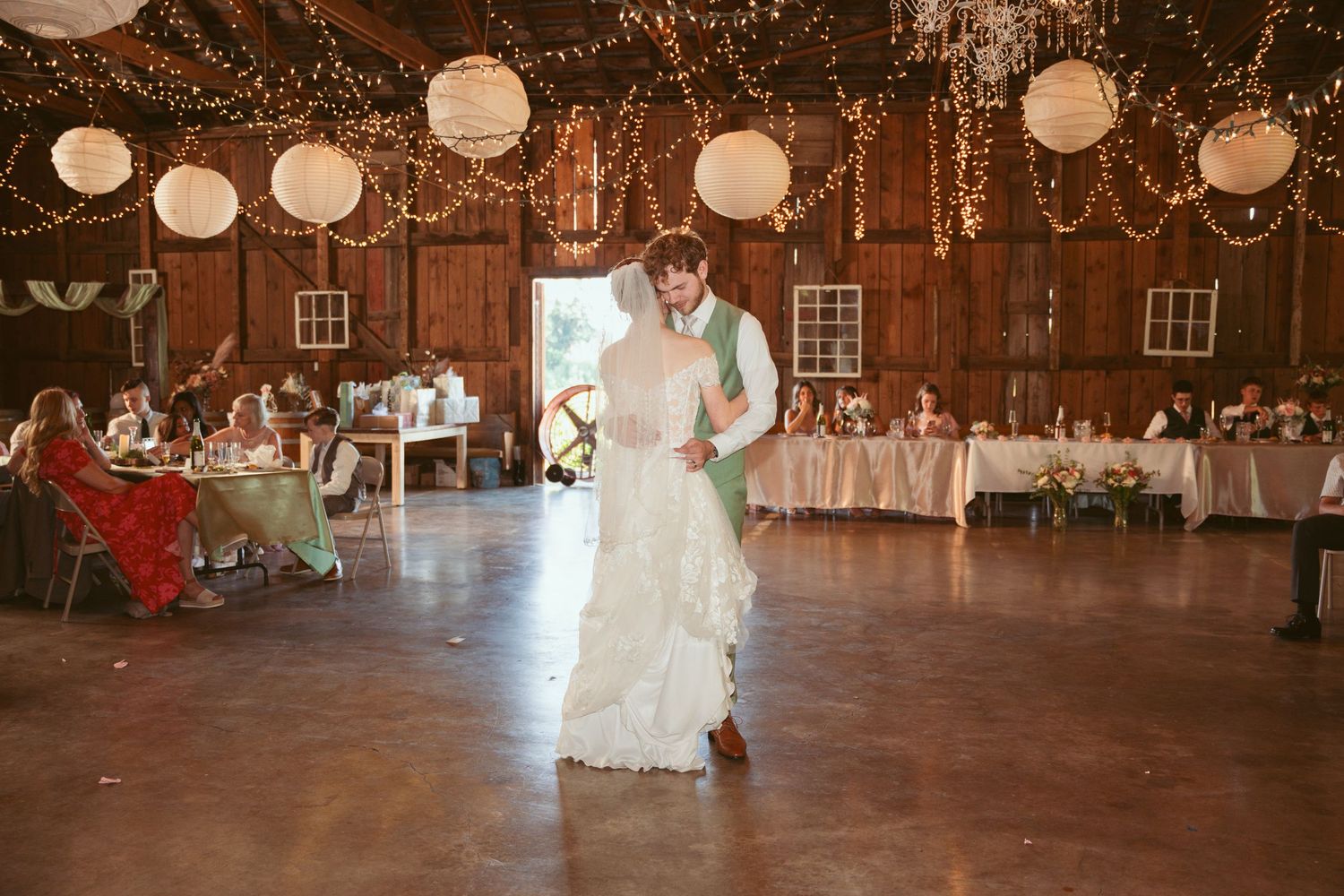 Wedding reception dance in a barn with twinkle lights and white paper lanterns overhead.