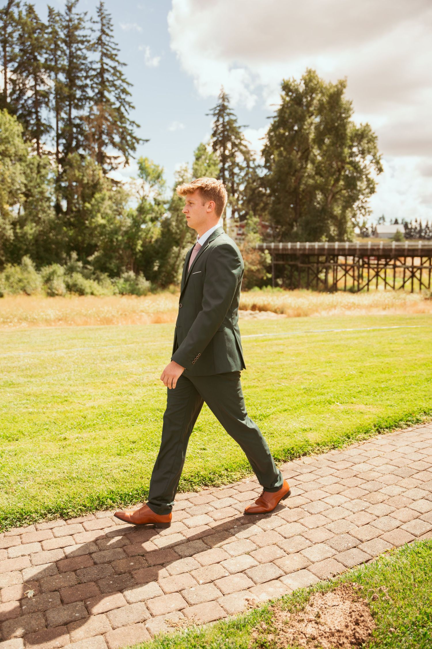 Person in formal attire walks along paved pathway with tall evergreen trees in background.