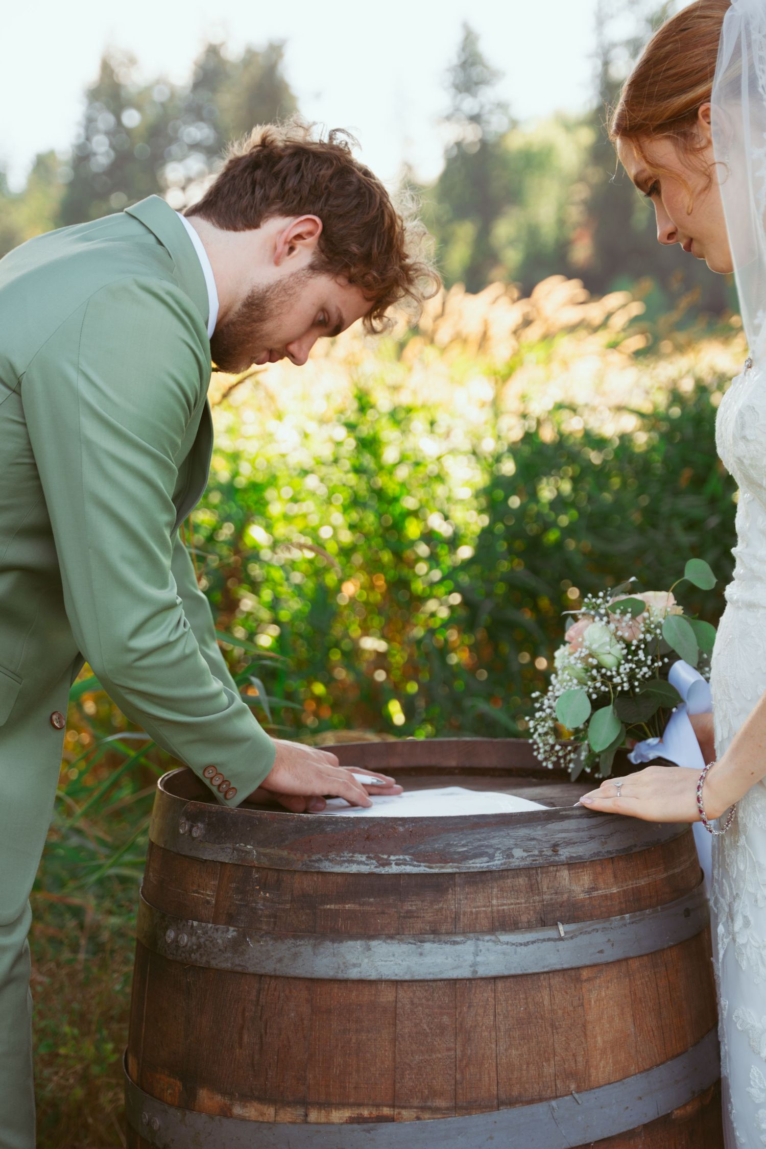 Wedding couple signs marriage documents on rustic wine barrel in outdoor garden setting.