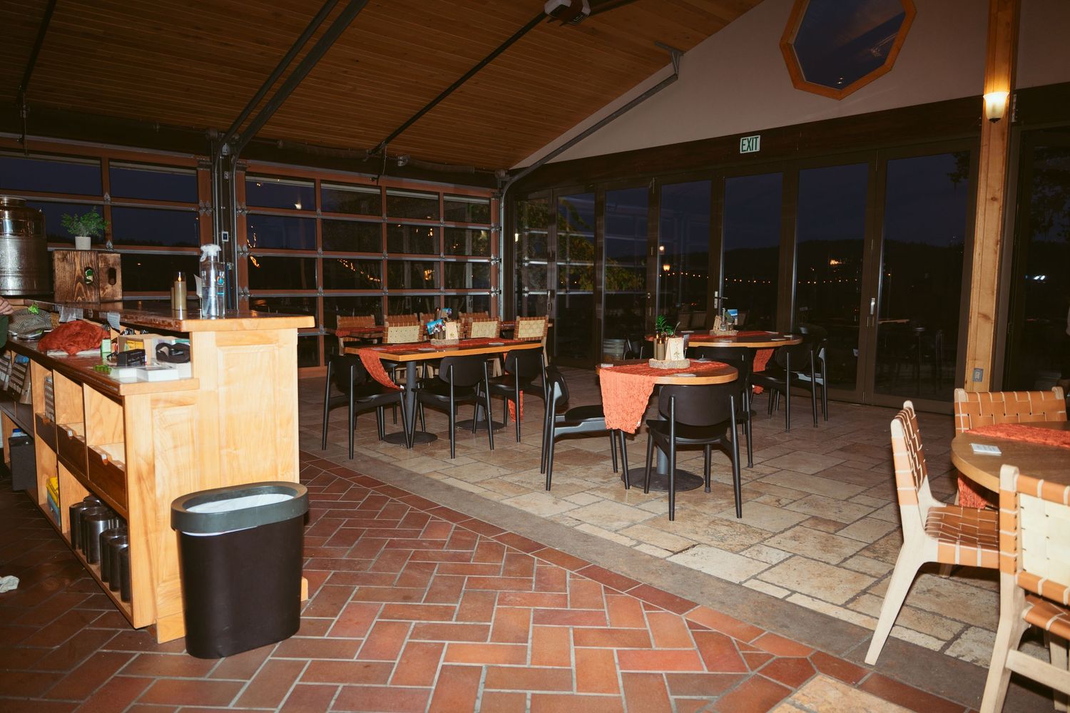 Restaurant interior with brick floor, wooden tables, and large windows at dusk.