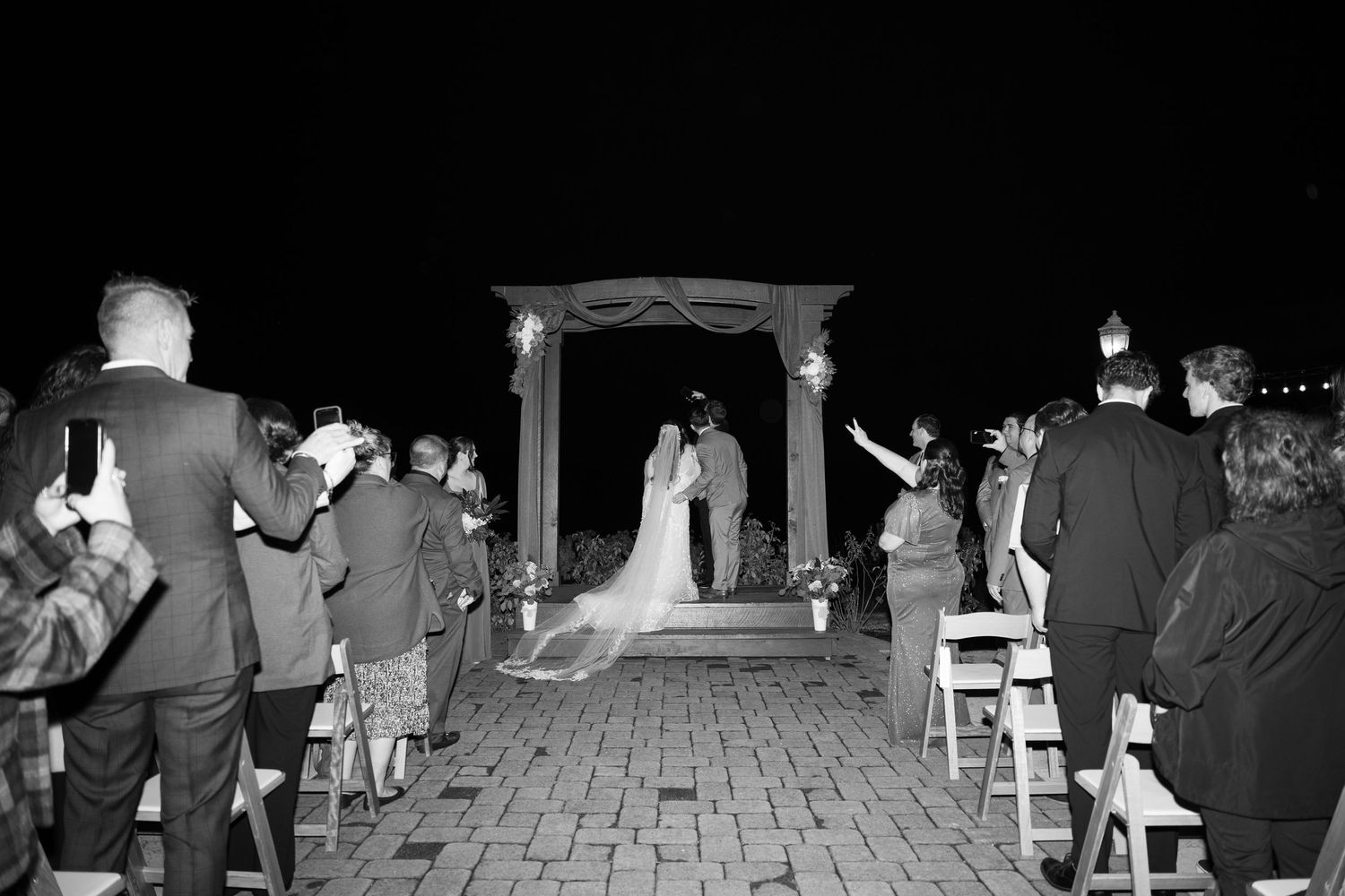 Nighttime outdoor wedding ceremony with guests seated in wooden chairs under twinkling lights.