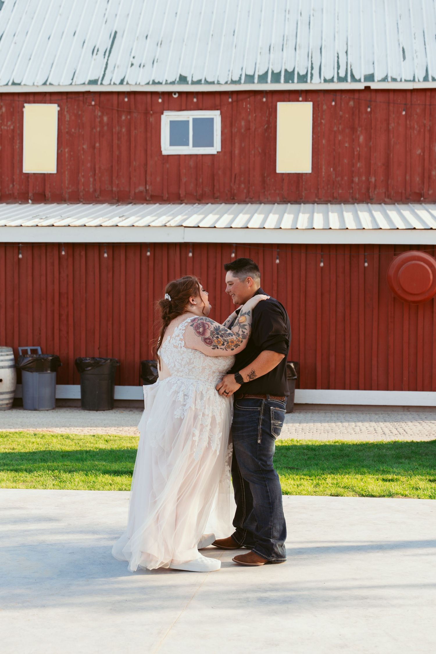 A wedding couple performs a playful dip dance move on a paved outdoor area near a red barn.