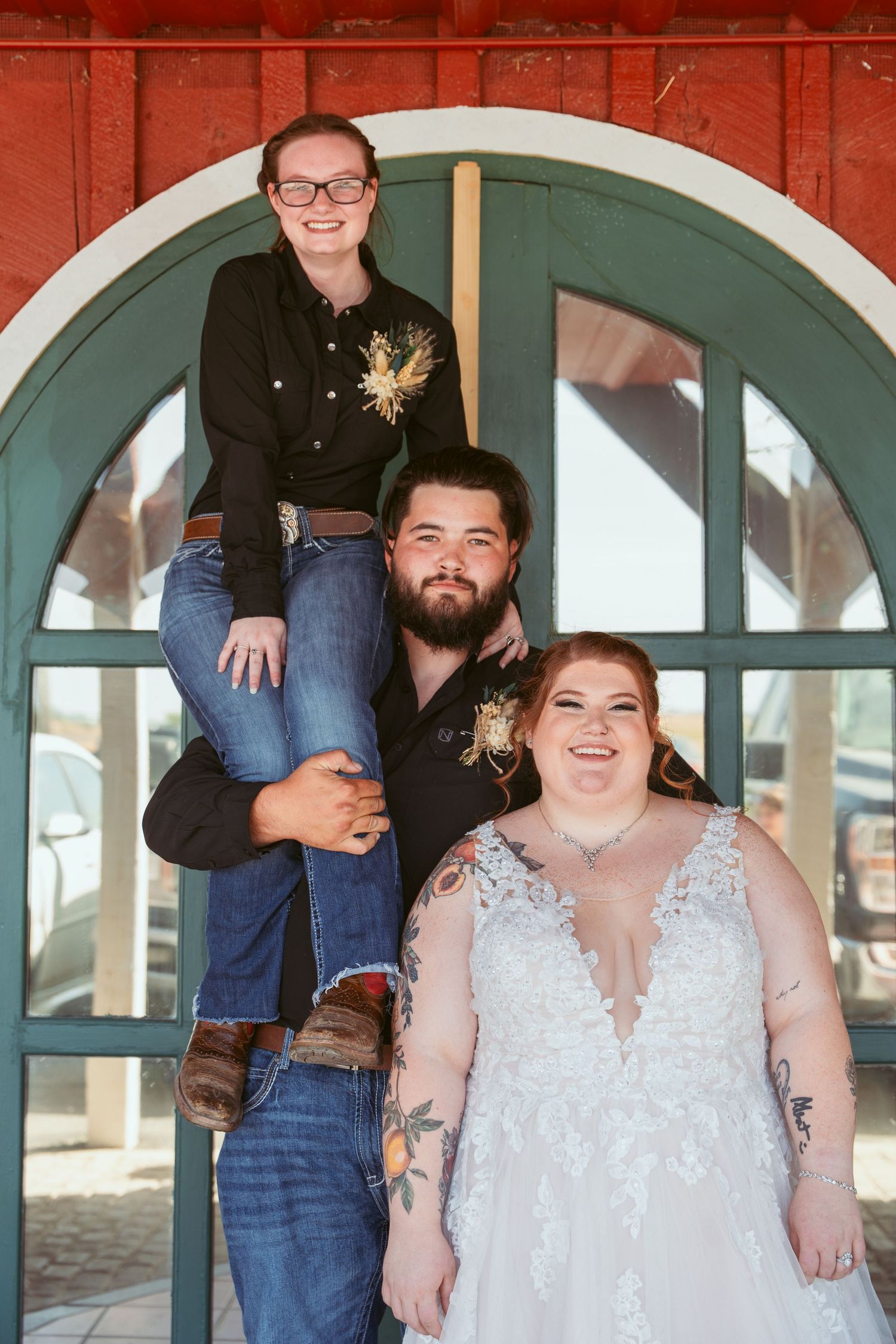 Wedding party members gather in front of rustic red barn with teal-trimmed archways.