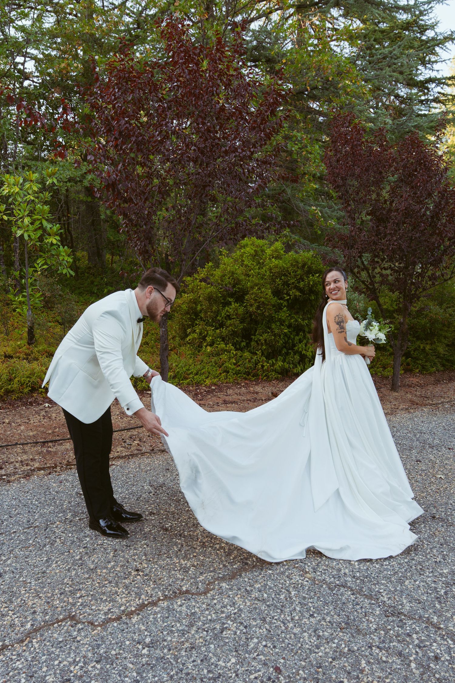 A wedding couple walks along a gravel path with trees and greenery in the background.