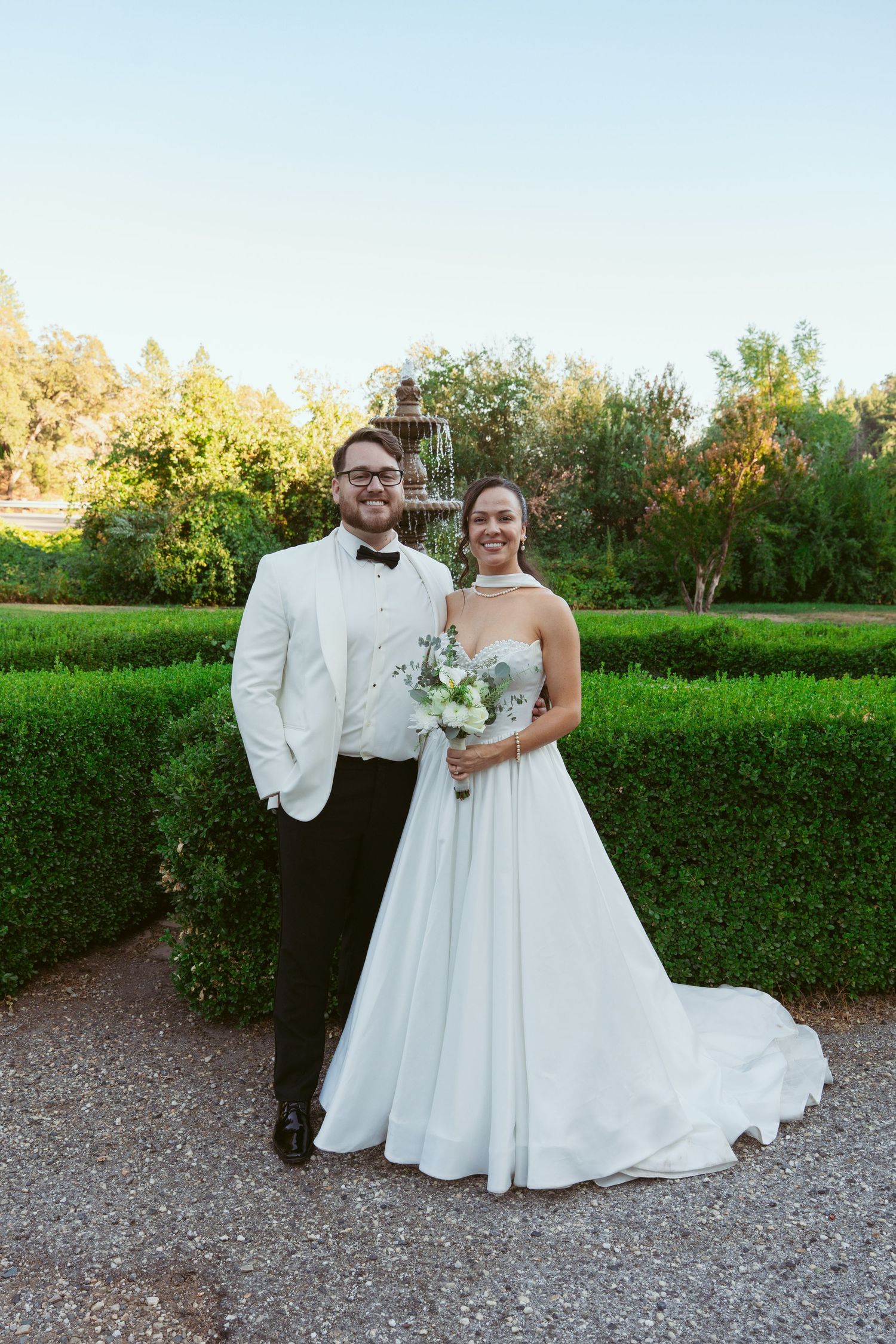 A wedding couple poses in formal attire against a garden backdrop with manicured hedges.