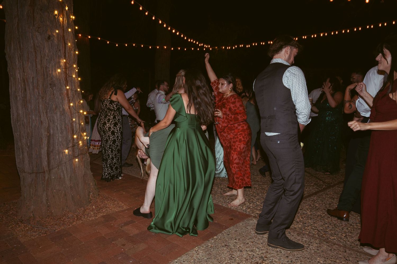 Outdoor wedding celebration with string lights and guests dancing in formal attire.