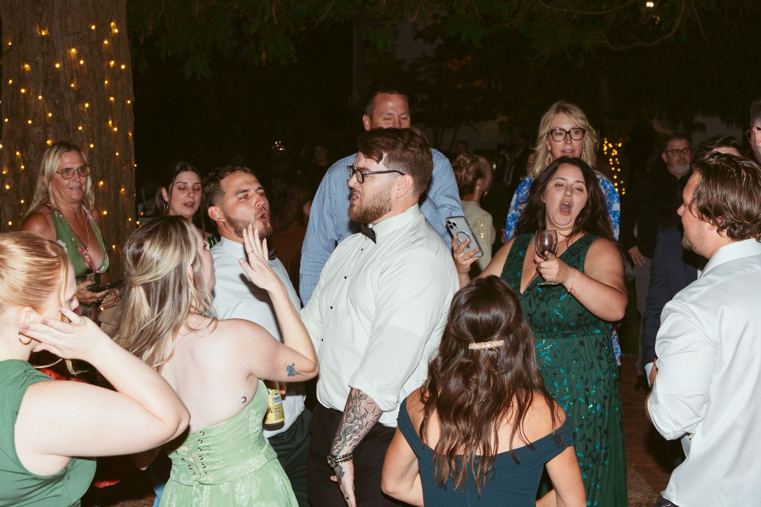 Wedding guests dance together under string lights during an evening reception celebration.