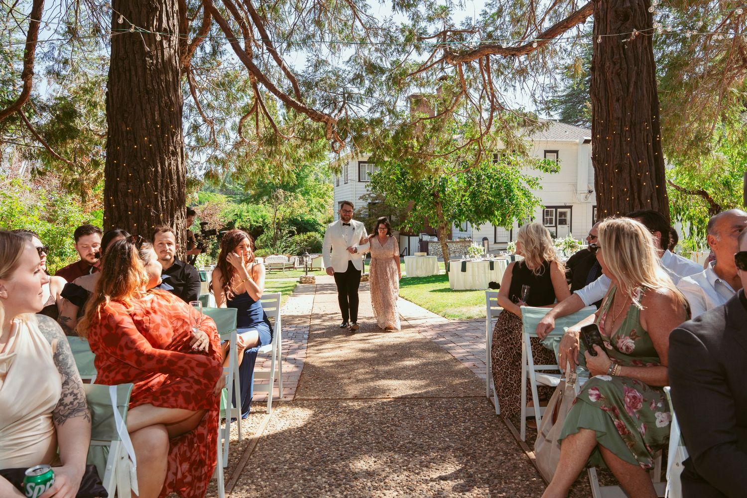 Wedding guests mingle and celebrate at an outdoor garden reception under tall trees.