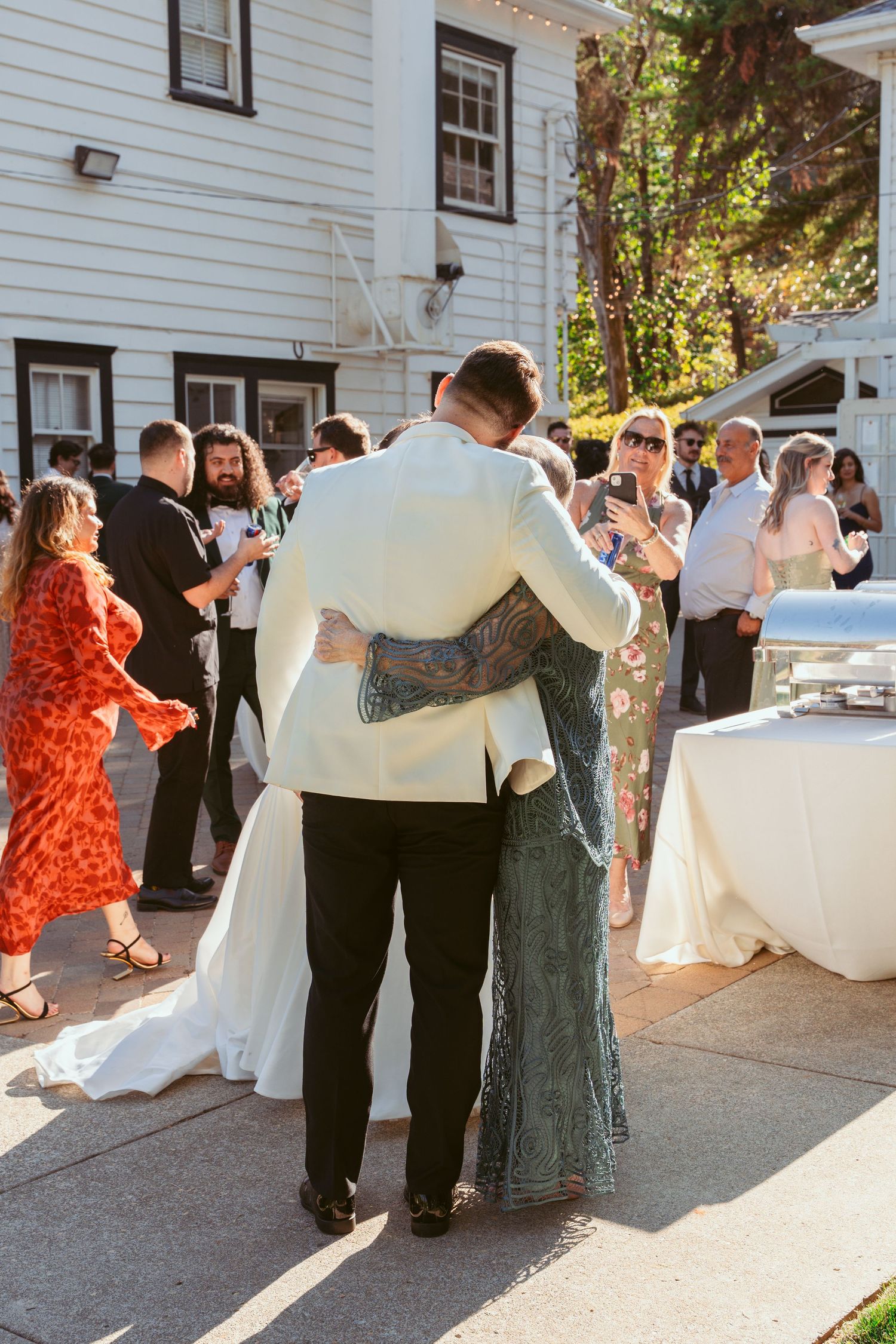 Wedding guests dance and celebrate at an outdoor reception near a white building.
