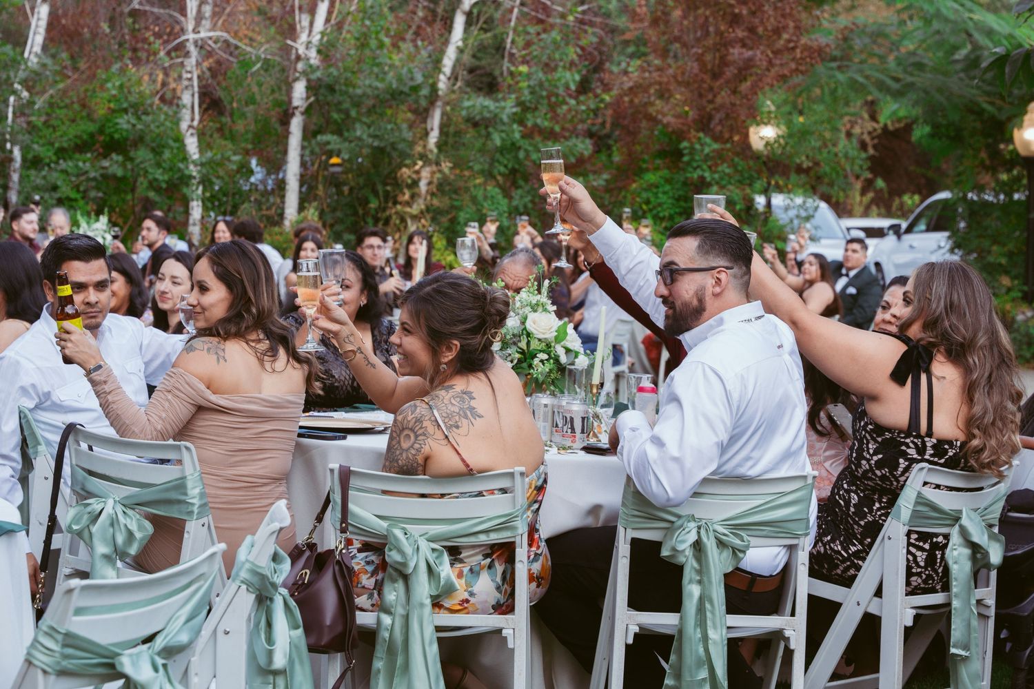 Wedding guests raise their glasses in a toast while seated at outdoor reception tables.