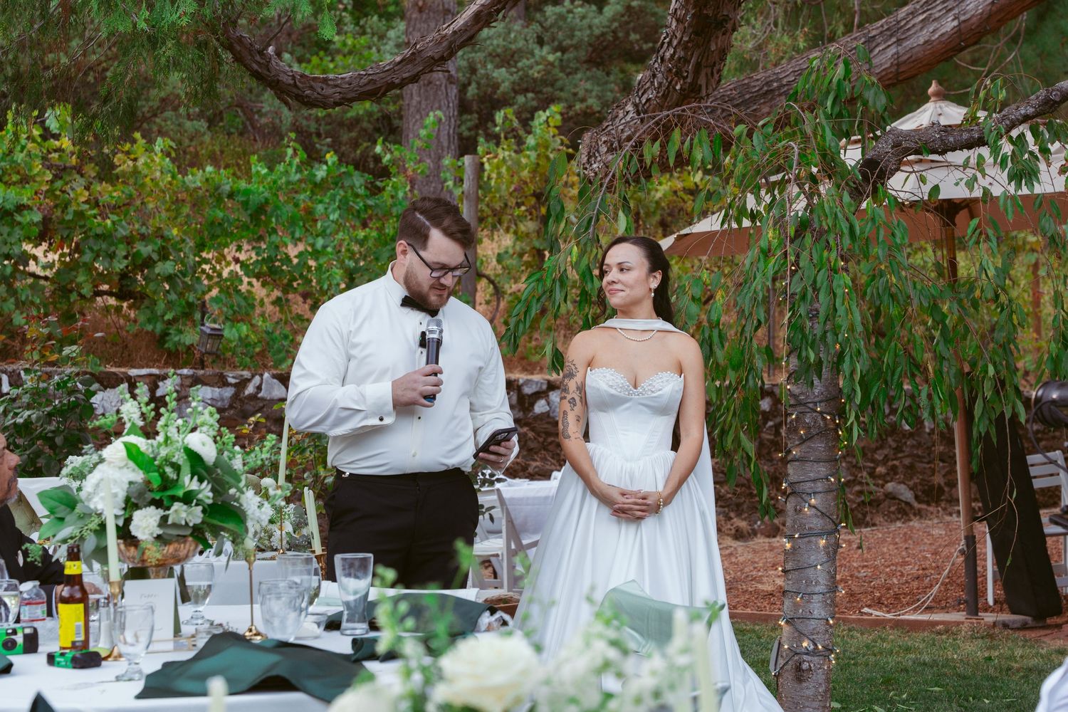 Wedding reception tables set outdoors with greenery and white decor under natural tree canopy.